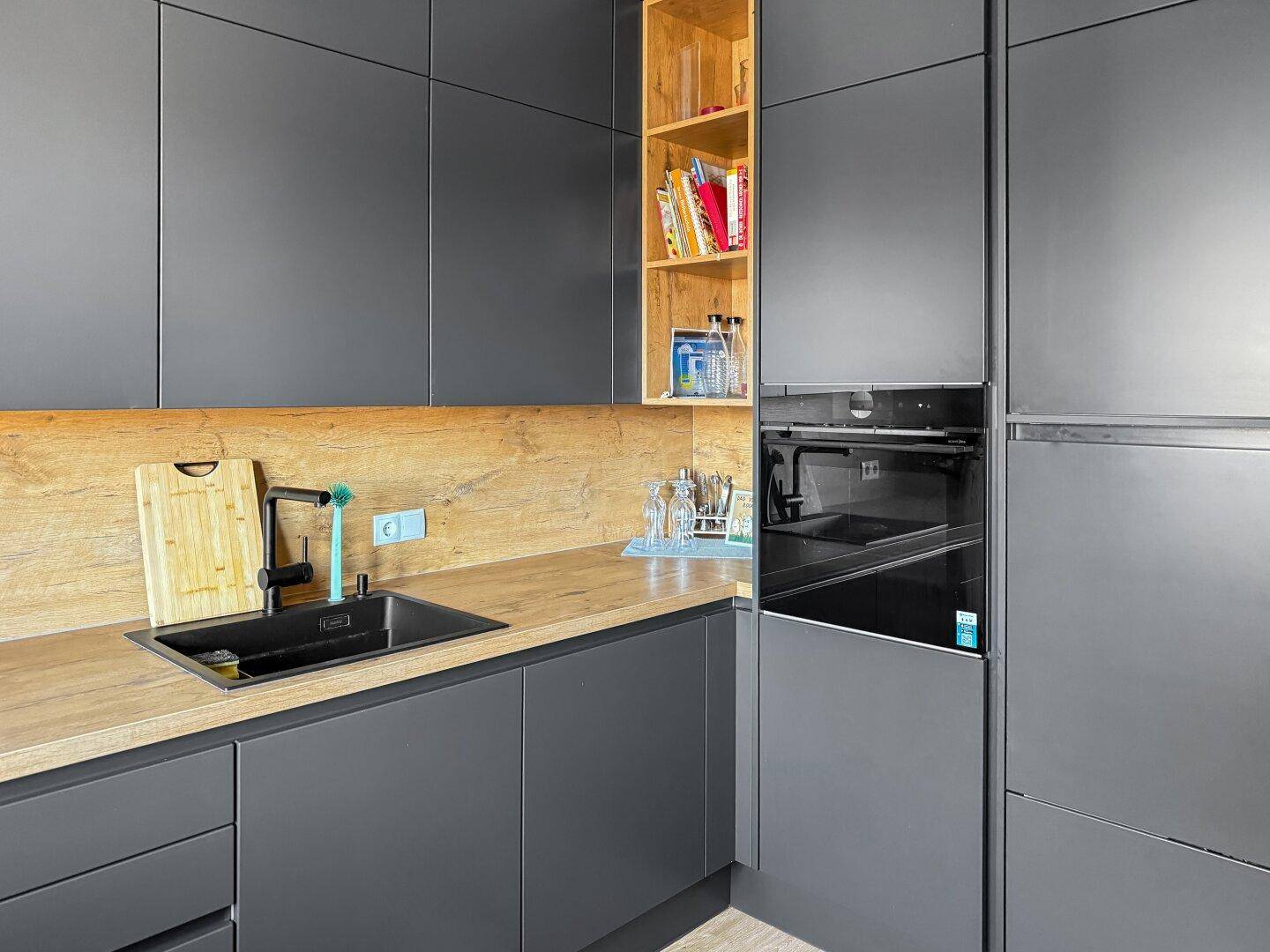 Integrierte Küche mit Backofen, dunklen Fronten und offenem Holzregal für Kochbücher.
