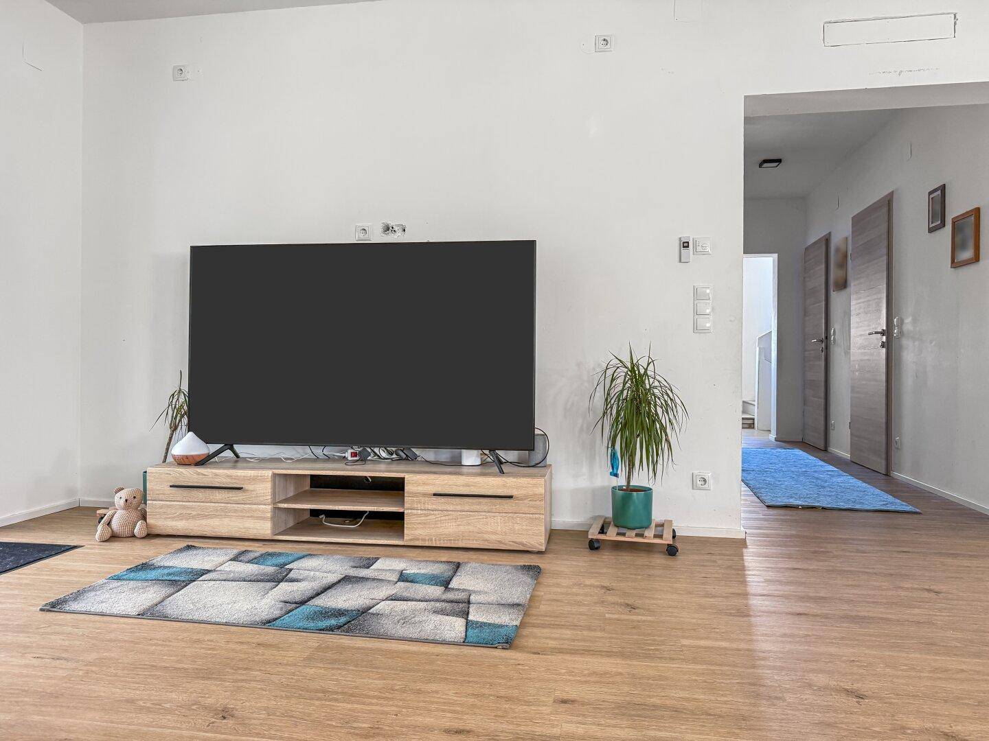 Heller Wohnbereich mit großem Fernseher, TV-Möbel und Zugang zu weiteren Räumen.