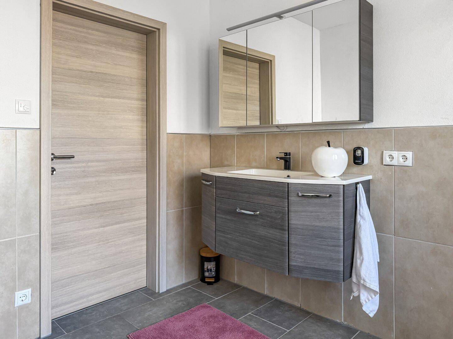Helles Badezimmer mit modernem Waschtisch, Spiegelschrank und Holztür.