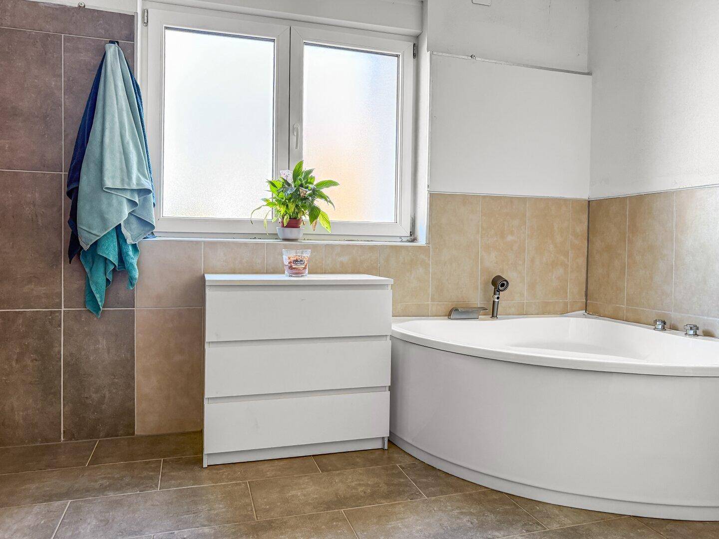 Badezimmer mit Eckbadewanne, Fenster und beigefarbenen Fliesen.