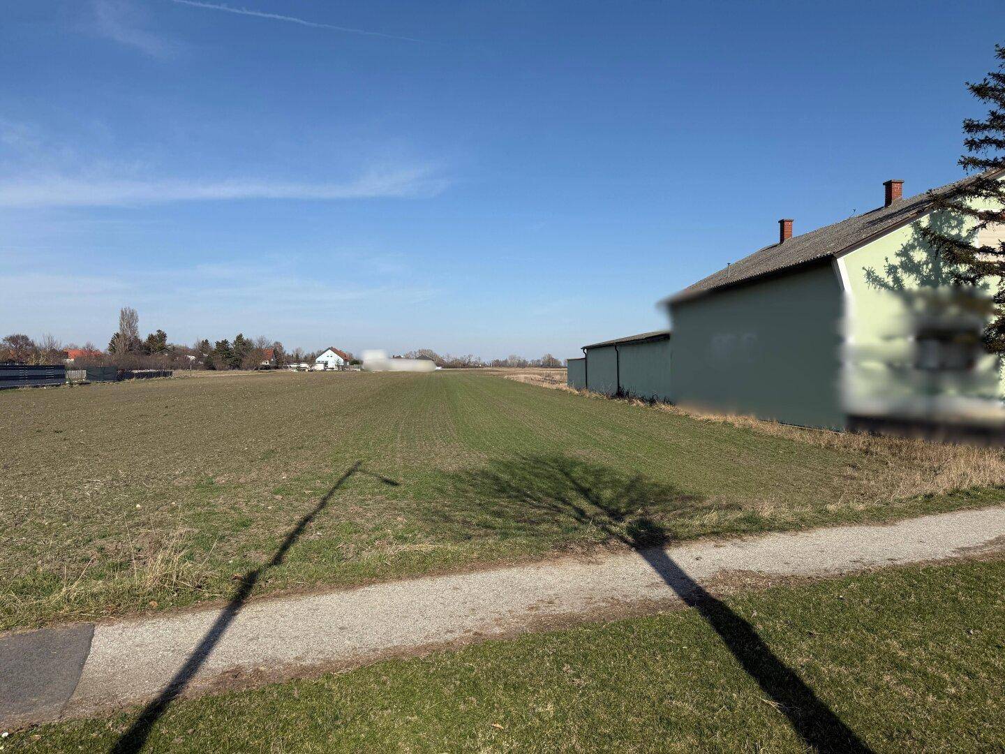 Blick über ein grünes Feld neben einem Weg, mit einem Gebäude rechts und Häusern in der Ferne.