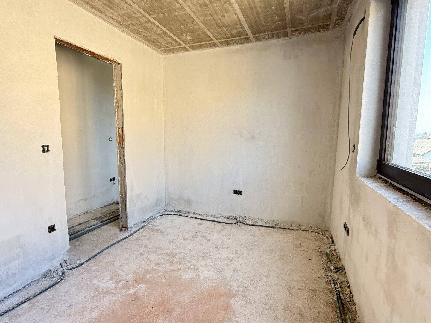 Rohbau-Zimmer mit Fenster, Betonboden und unfertigen Wänden.