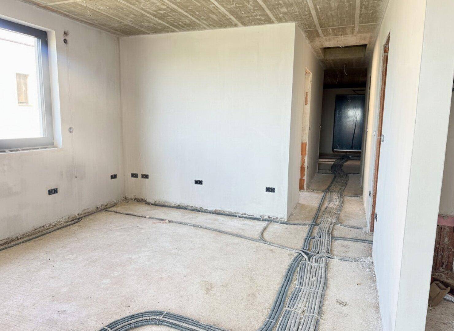 Rohbau-Raum mit Fenster, Betonboden und sichtbaren Elektroinstallationen.