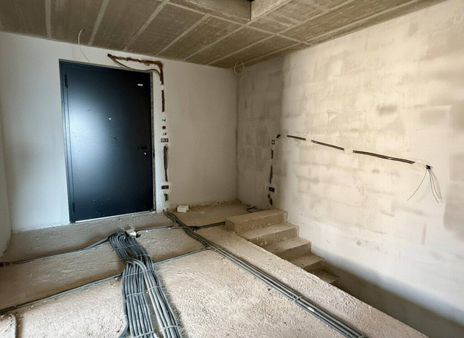 Eingangsbereich im Rohbau mit schwarzer Tür und sichtbaren Elektroinstallationen.