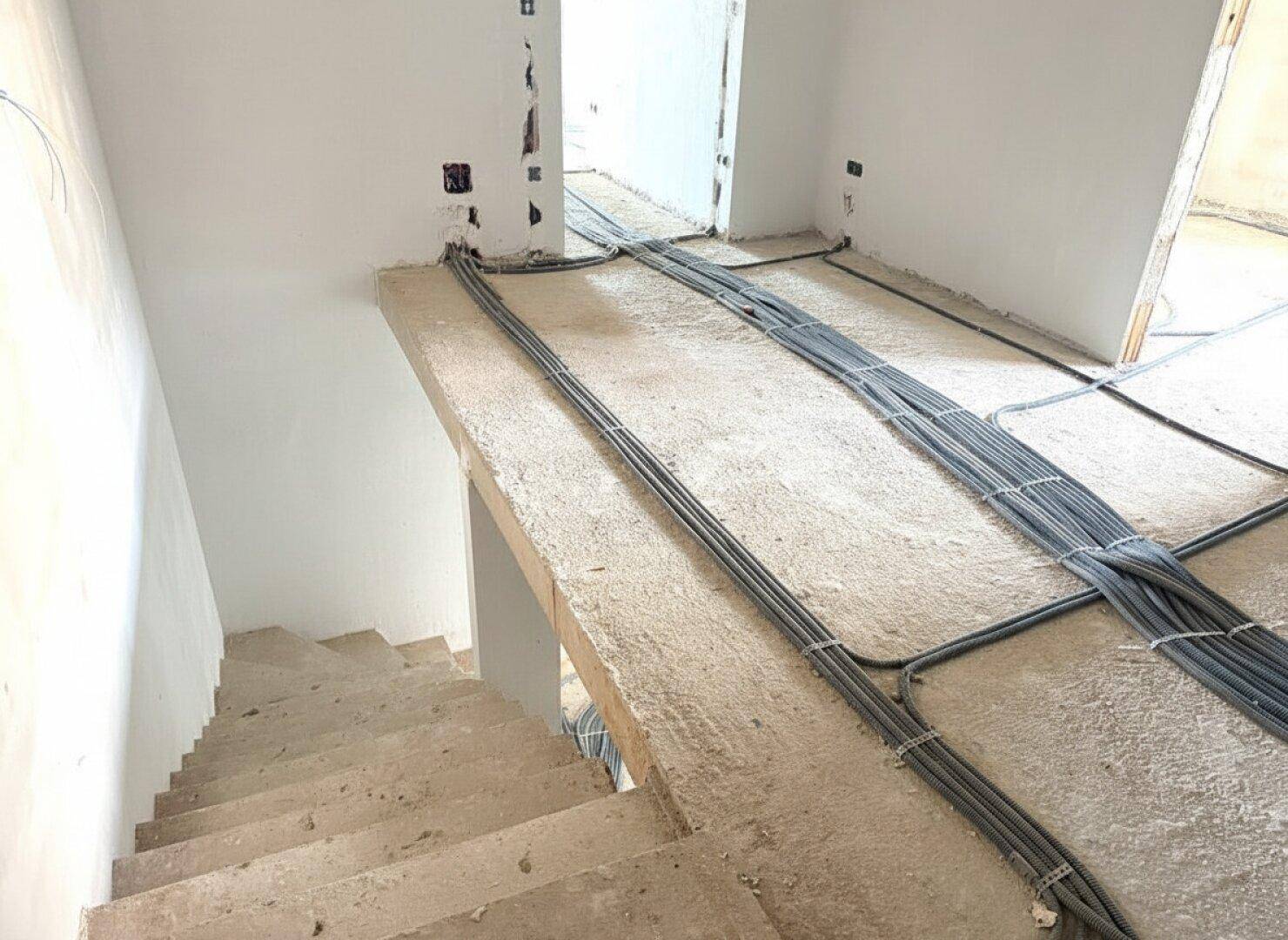 Flur im Rohbau mit Treppenaufgang und sichtbaren Elektroinstallationen.