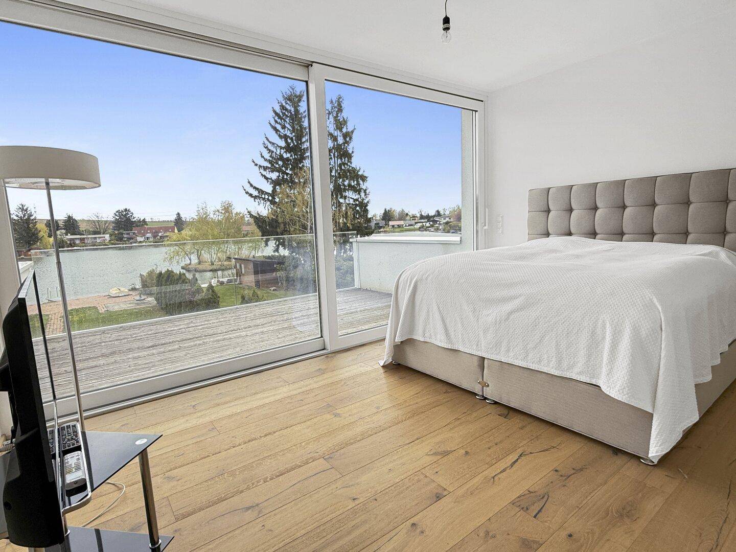 Schlafzimmer mit Holzboden, großen Fenstern und direktem Blick auf den See.