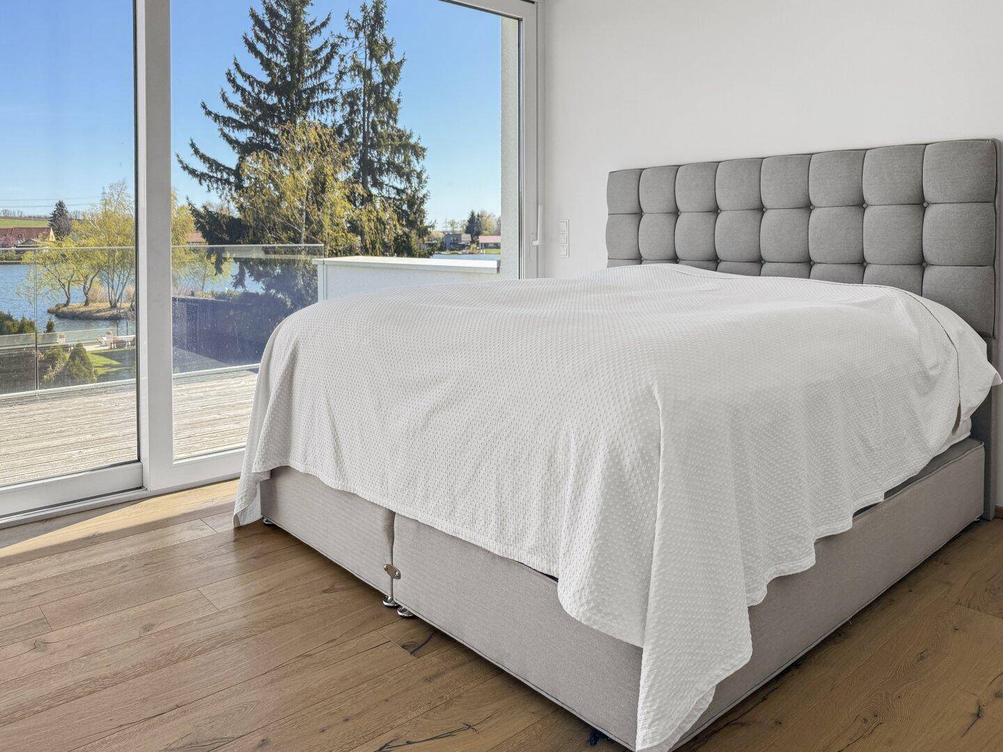 Schlafzimmer mit Holzboden, großen Fenstern und direktem Blick auf den See.