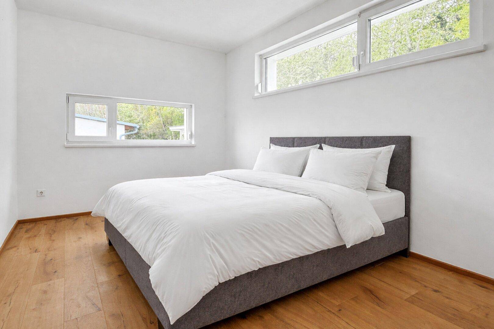 Schlafzimmer mit Holzboden, weißen Wänden und zwei Fenstern für viel Tageslicht.