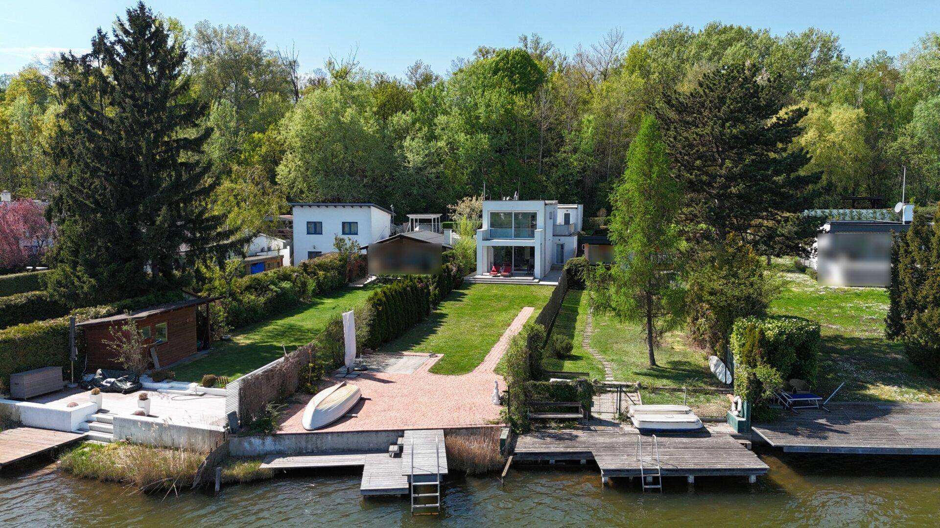 Luftaufnahme des modernen Hauses am Seeufer mit großem Garten und Bootsstegen.