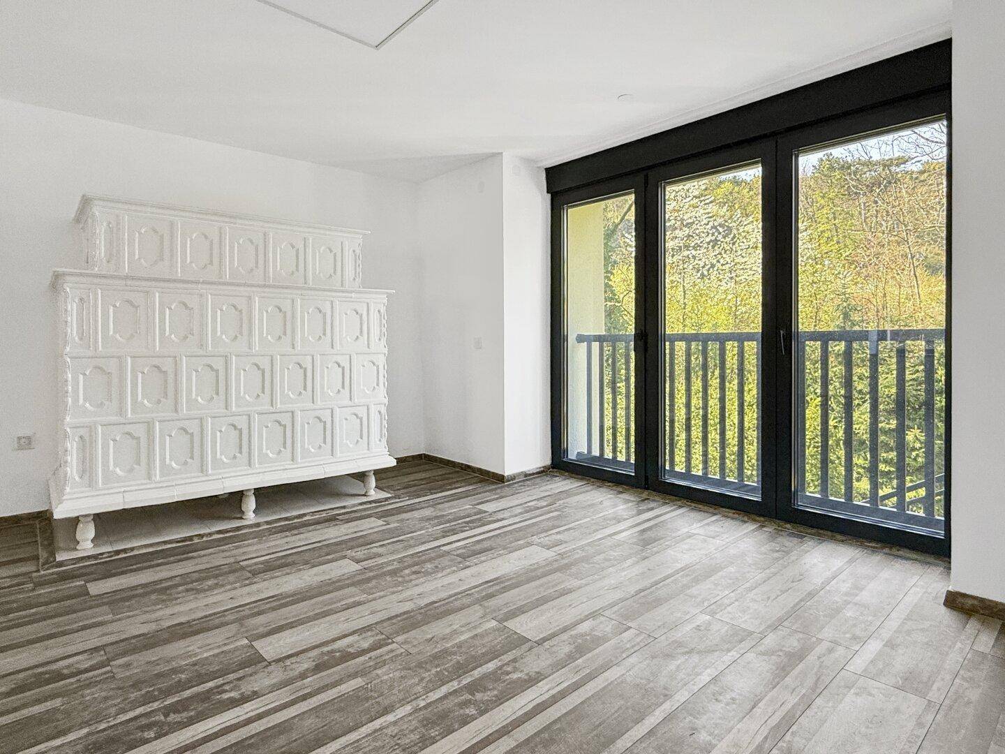 Heller Raum mit traditionellem Kachelofen, Holzbodenoptik und Zugang zum Balkon.
