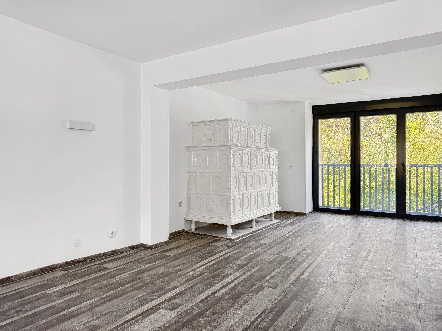 Heller Wohnraum mit Kachelofen und großen Fenstern zum Balkon.