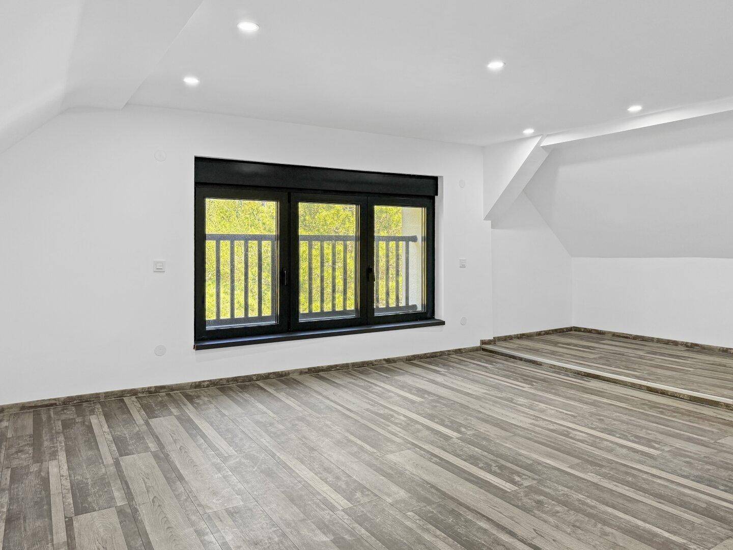 Heller Raum im Dachgeschoss mit Holzbodenoptik, großen Fenstern und Deckenspots.