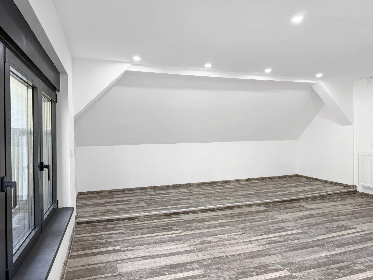 Geräumiger Raum im Dachgeschoss mit Holzbodenoptik, Deckenspots und schrägen Wänden.