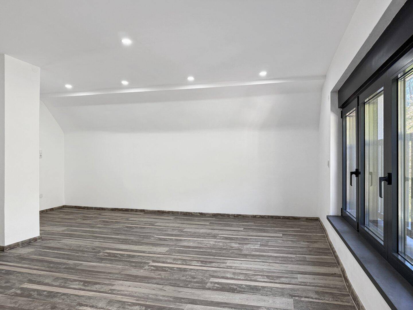 Heller Raum im Dachgeschoss mit Holzbodenoptik, großen Fenstern und Deckenspots.