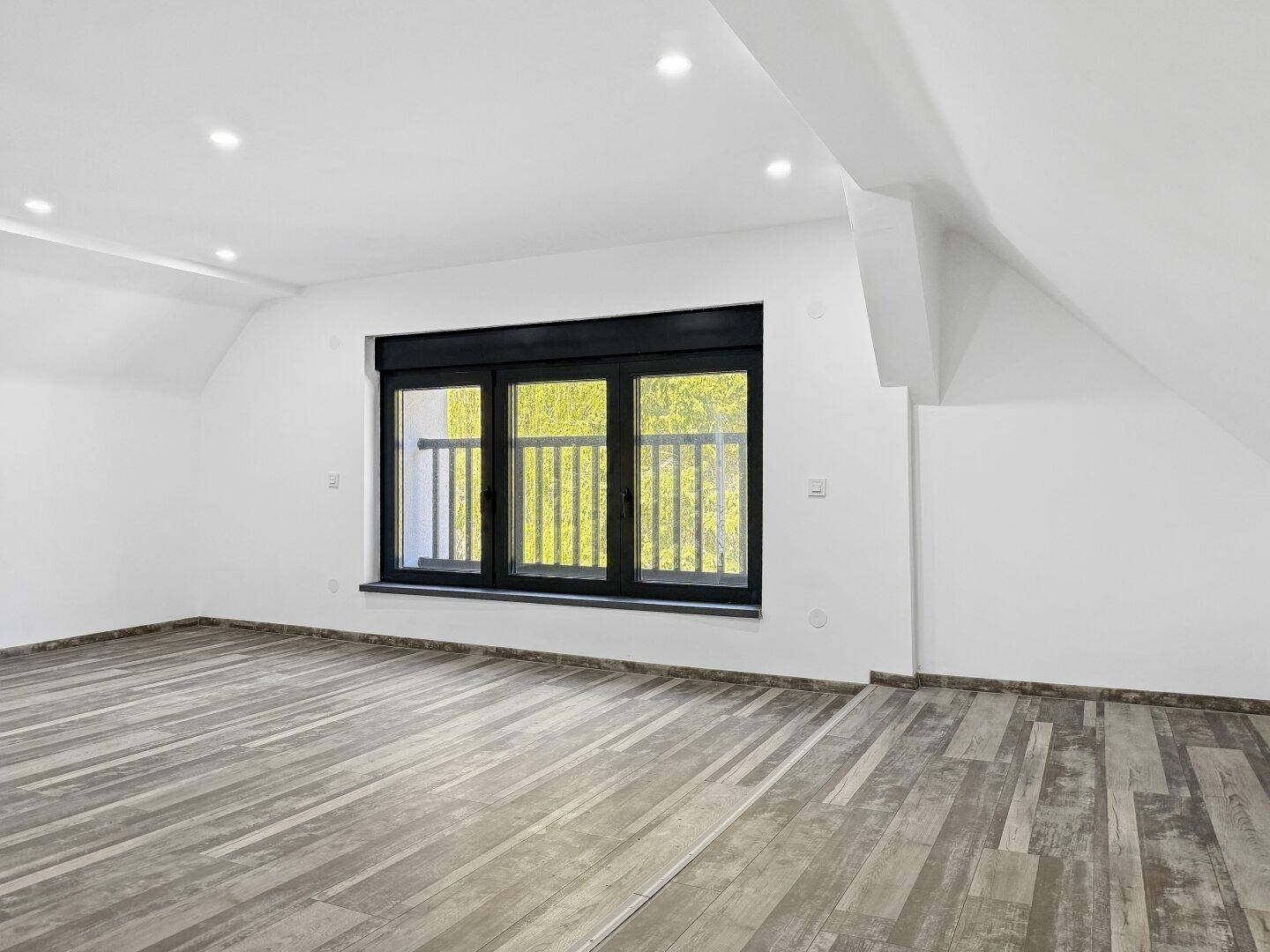 Heller Raum im Dachgeschoss mit Holzbodenoptik, großen Fenstern und Deckenspots.