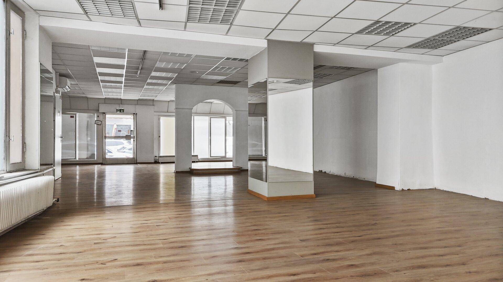 Großer leerer Gewerberaum mit Laminatboden, Rasterdecke und großen Fenstern.