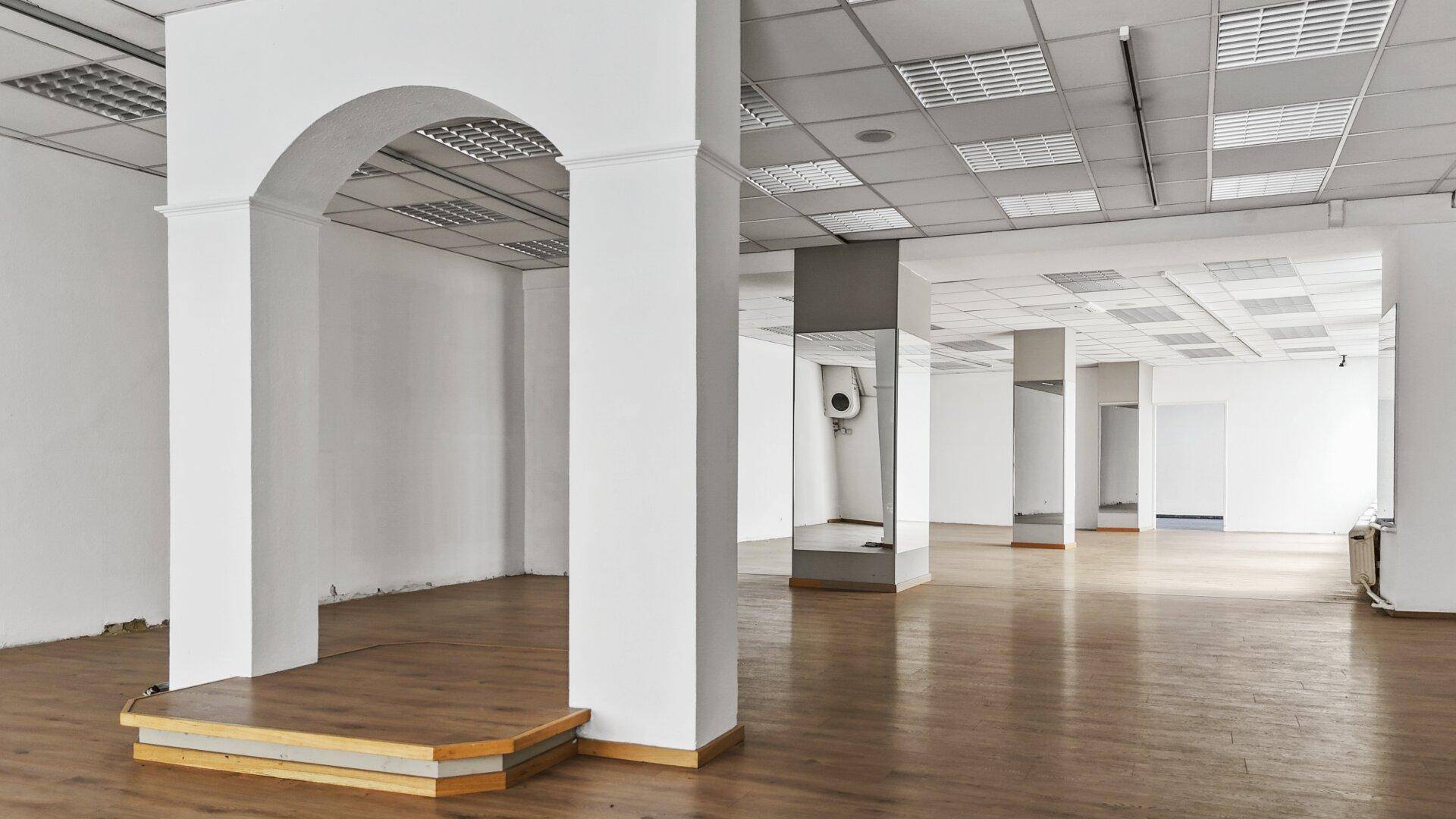 Geräumiger Gewerberaum mit Laminatboden, Rasterdecke, verspiegelten Säulen und einem Bogen.