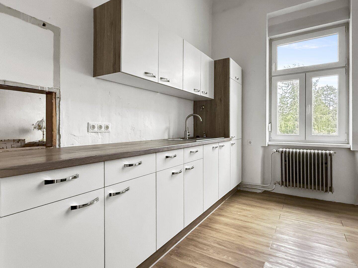 Einfache Küche mit weißen Schränken, Holzoptik-Arbeitsplatte und einem Fenster mit Blick ins Grüne.