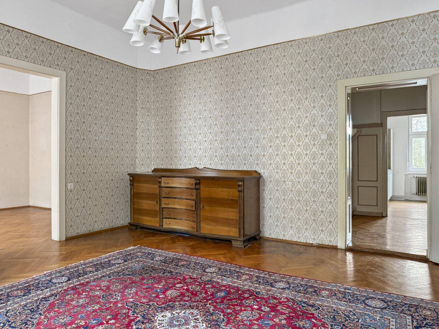 Zimmer mit Parkettboden und gemusterter Tapete, Blick auf ein Sideboard und offene Türen.