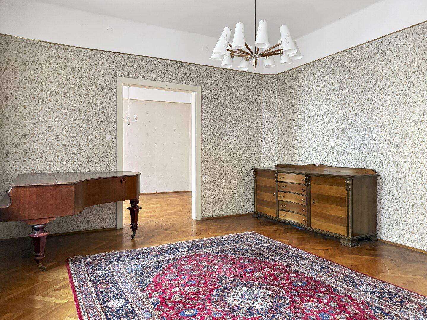 Geräumiges Zimmer mit Parkettboden, gemusterter Tapete und einem Flügel sowie einem Sideboard.