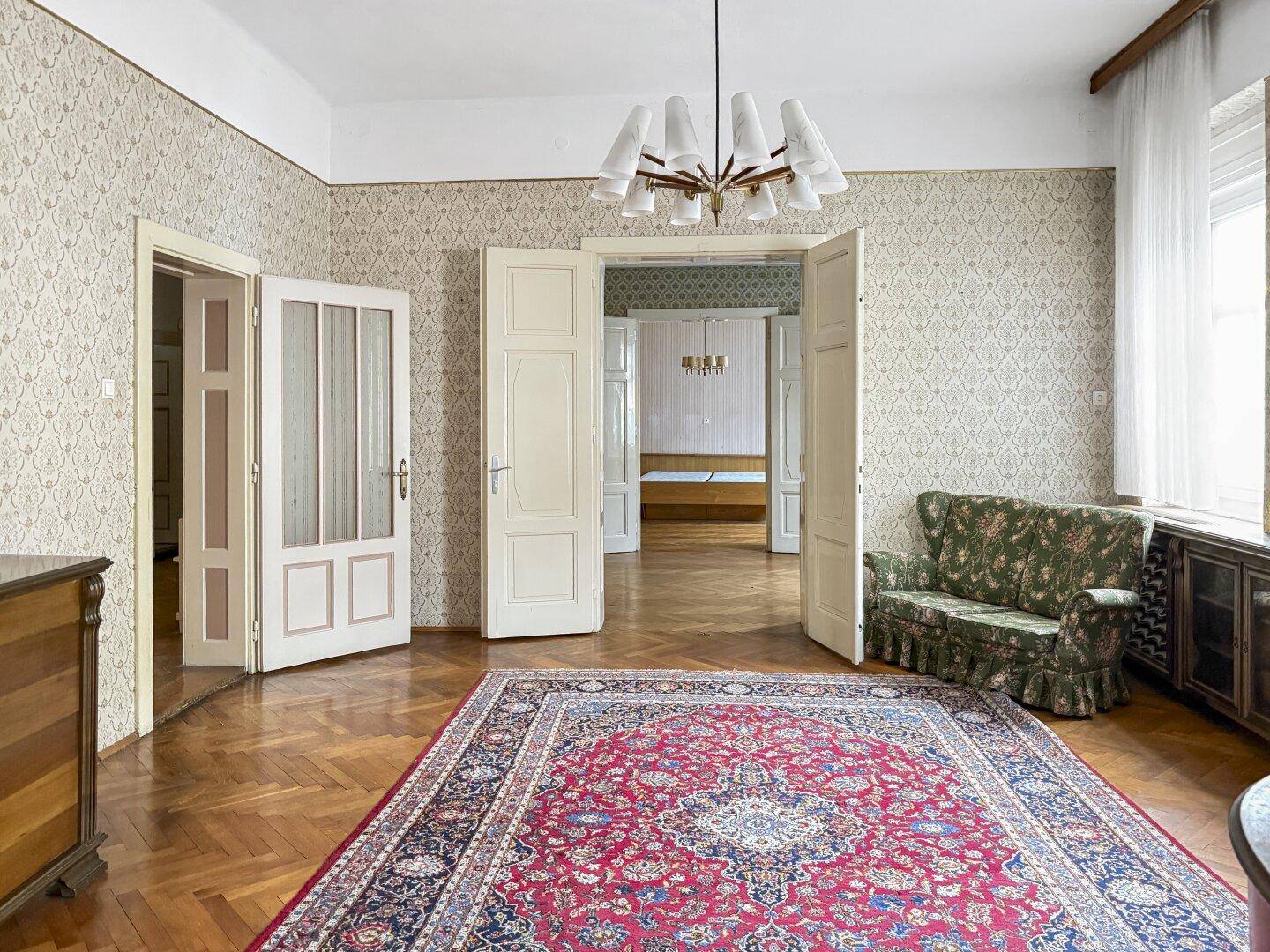 Großes Zimmer mit Fischgrätparkett, gemusterter Tapete und einem traditionellen Teppich.