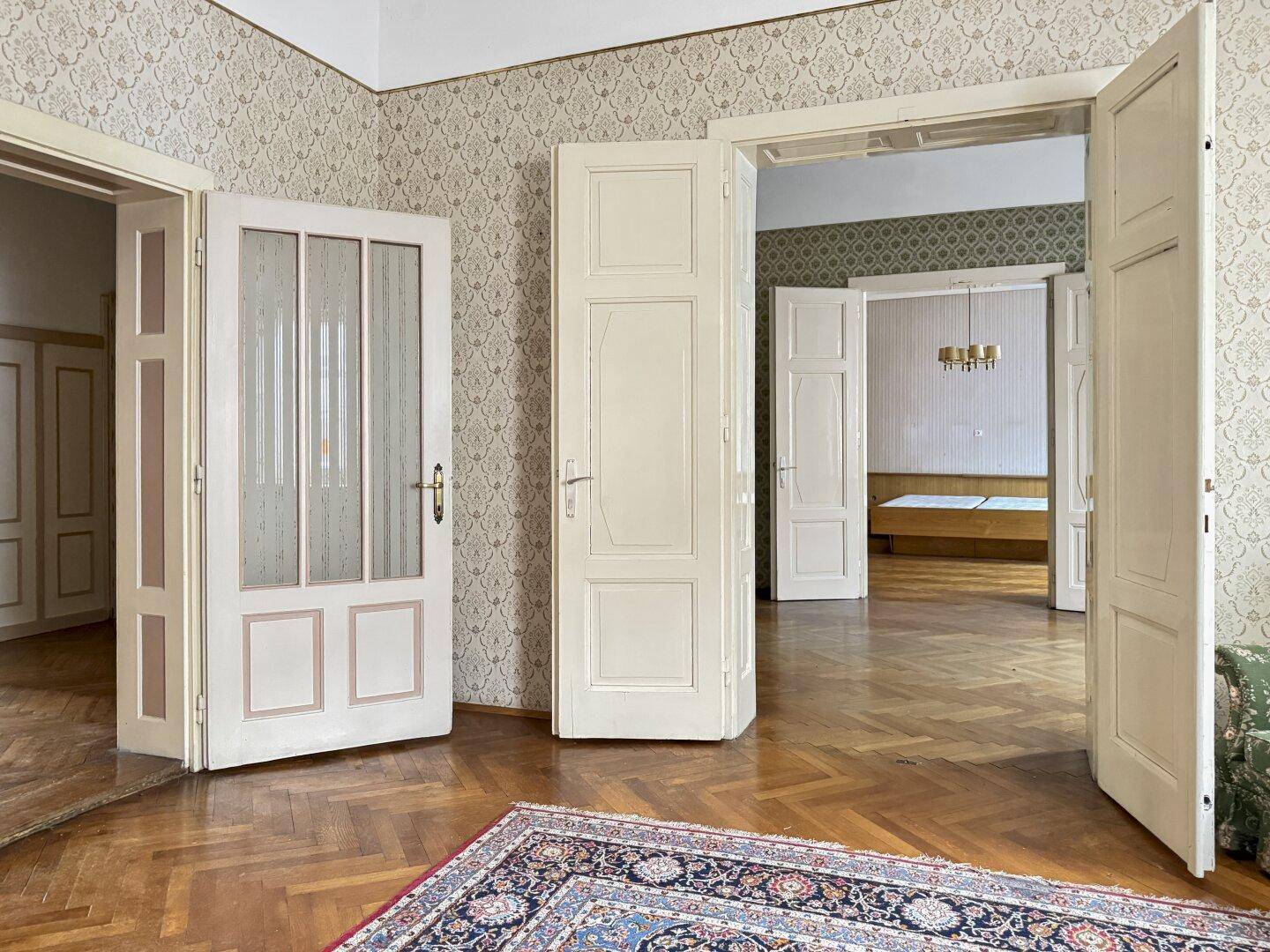 Zimmer mit Parkettboden und gemusterter Tapete, offene Doppeltüren führen in einen weiteren Raum.