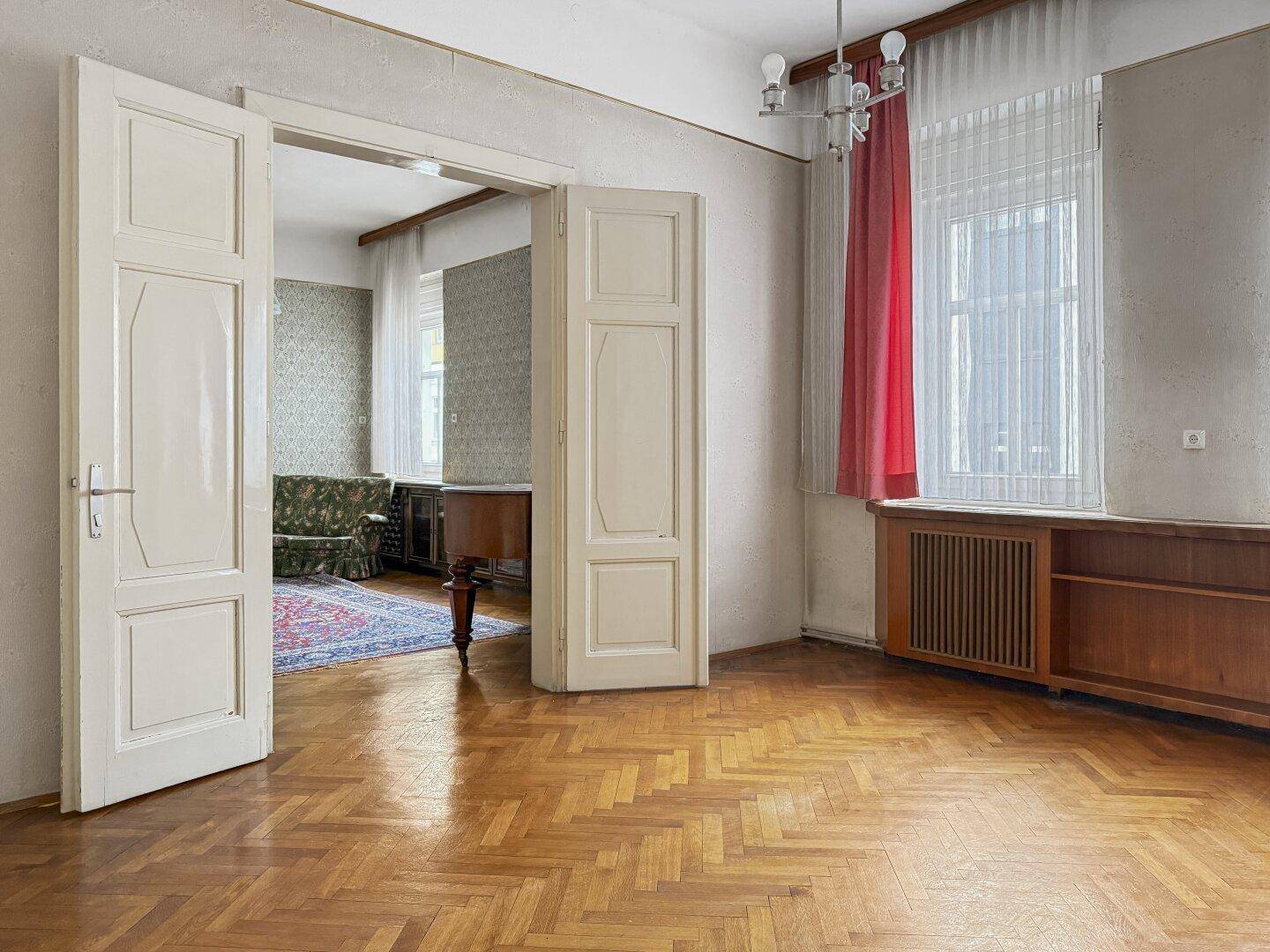Zimmer mit Parkettboden, gemusterter Tapete und einem Fenster mit roten Vorhängen.