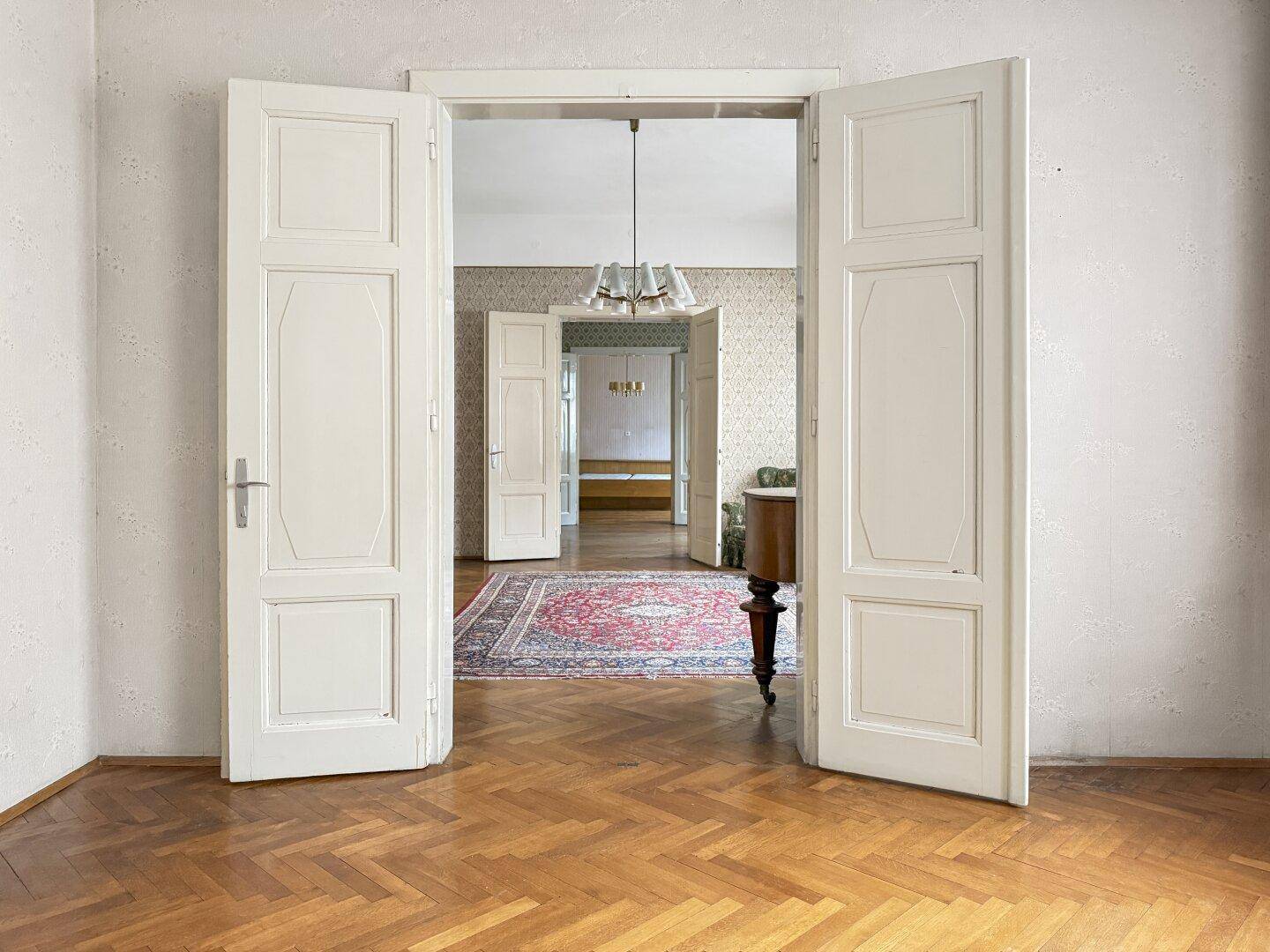 Zimmer mit Parkettboden und gemusterter Tapete, offene Doppeltüren führen in einen weiteren Raum.
