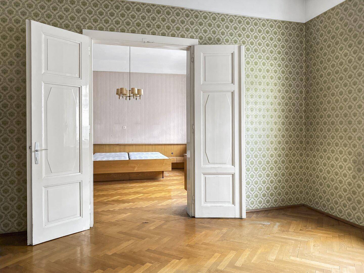 Zimmer mit Parkettboden und gemusterter Tapete, offene Doppeltüren führen in einen weiteren Raum.