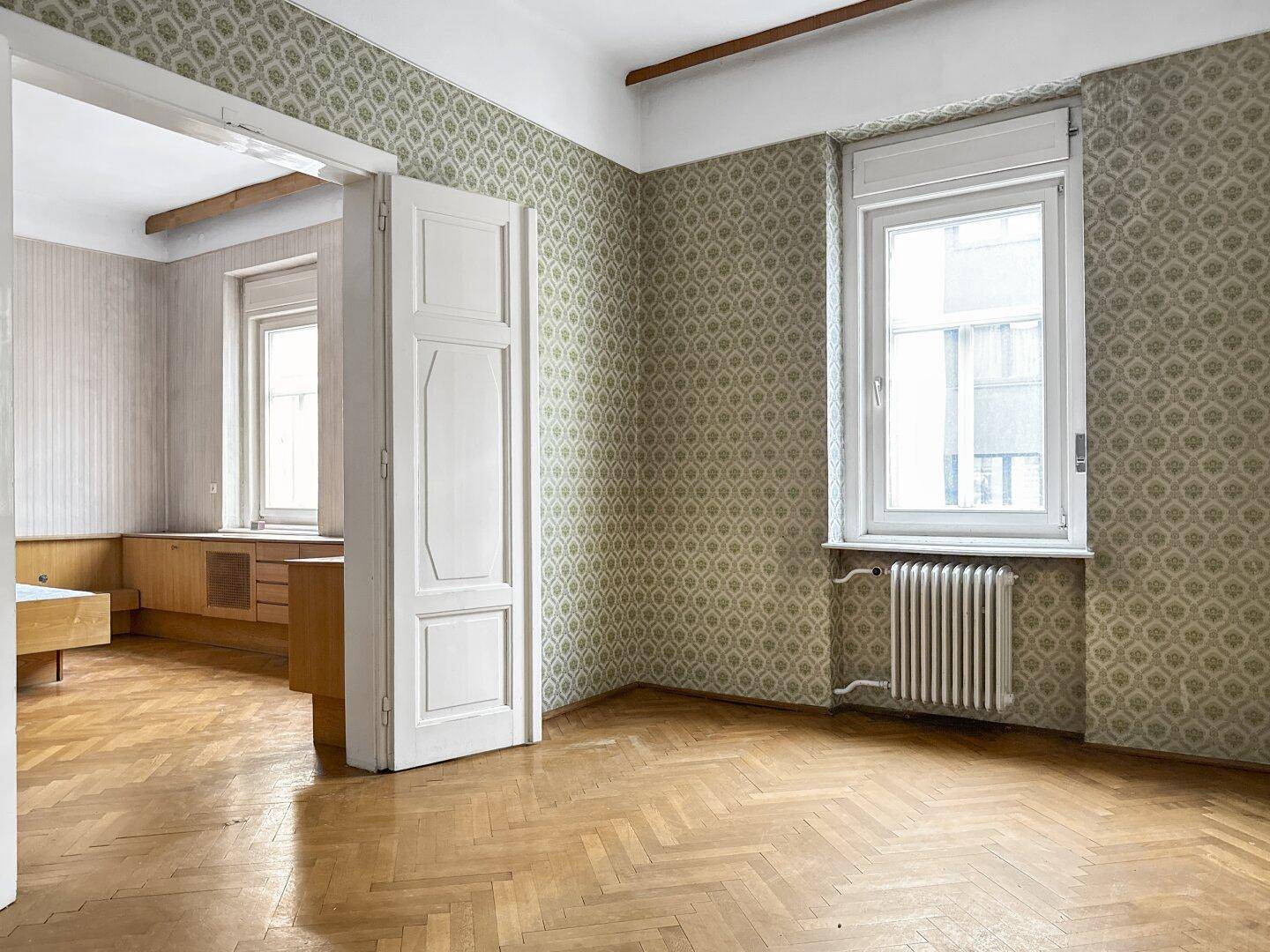 Zimmer mit Parkettboden, gemusterter Tapete und einem Fenster, das viel Tageslicht hereinlässt.