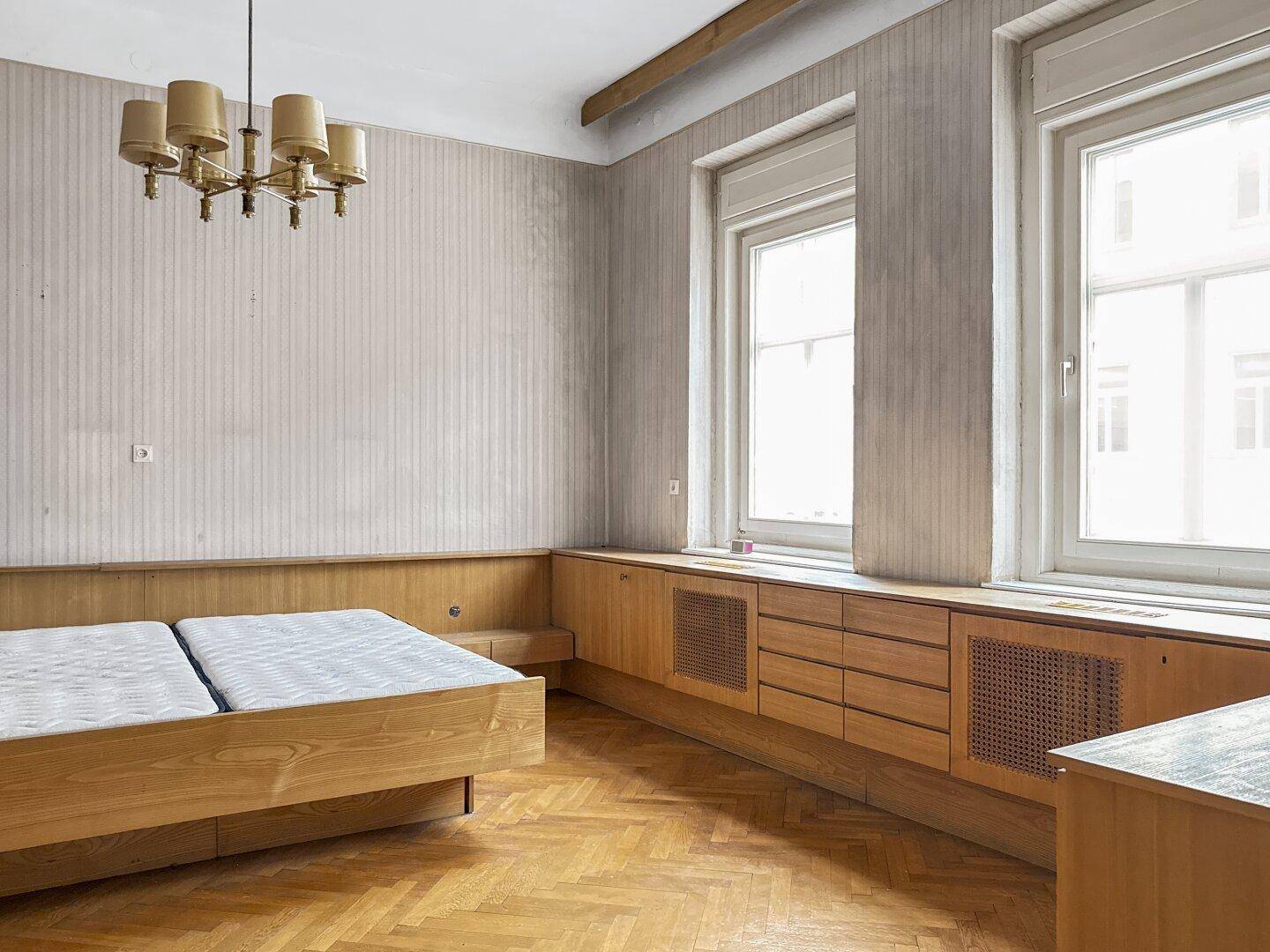 Schlafzimmer mit zwei Einzelbetten, Einbaumöbeln unter den Fenstern und Fischgrätparkett.