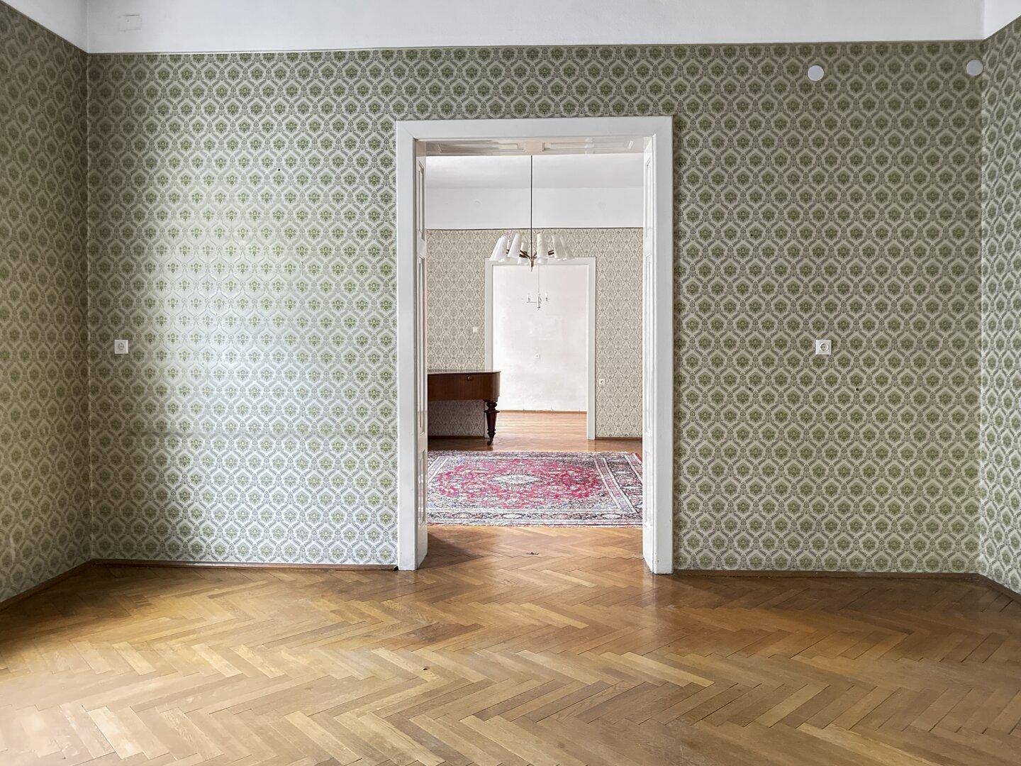 Leeres Zimmer mit gemusterter Tapete, Fischgrätparkett und Zugang zu einem weiteren Raum.