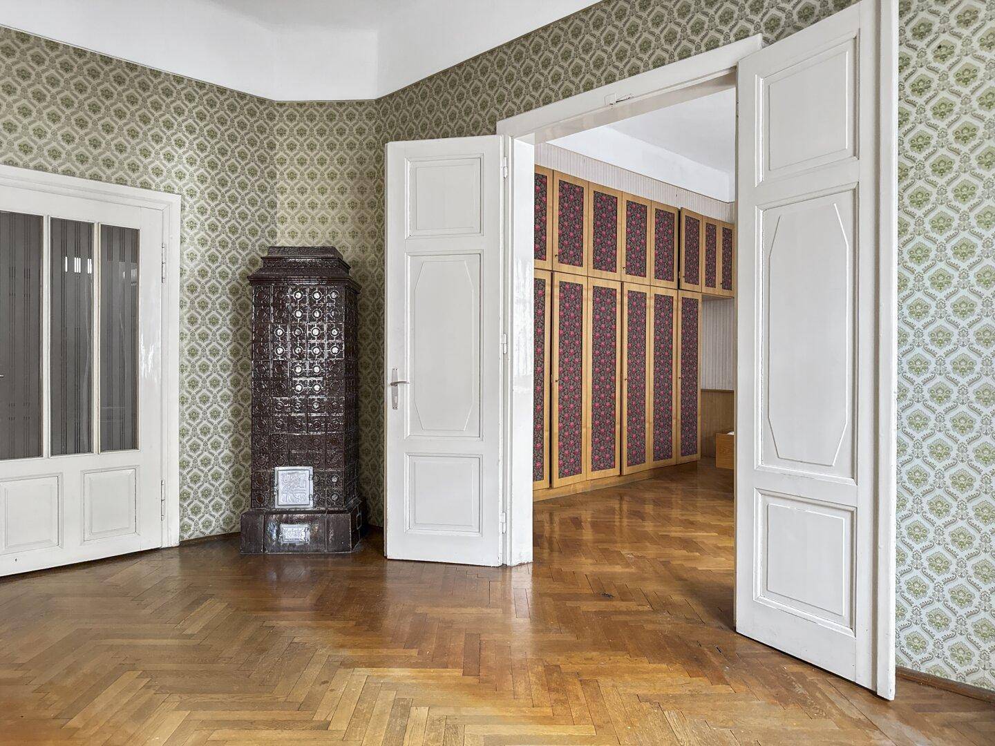 Geräumiges Zimmer mit Fischgrätparkett, gemusterter Tapete und einem traditionellen Kachelofen.