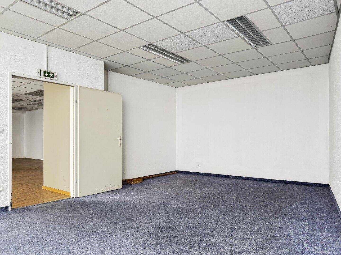Zimmer mit blauem Teppichboden, weißen Wänden und einer Rasterdecke.