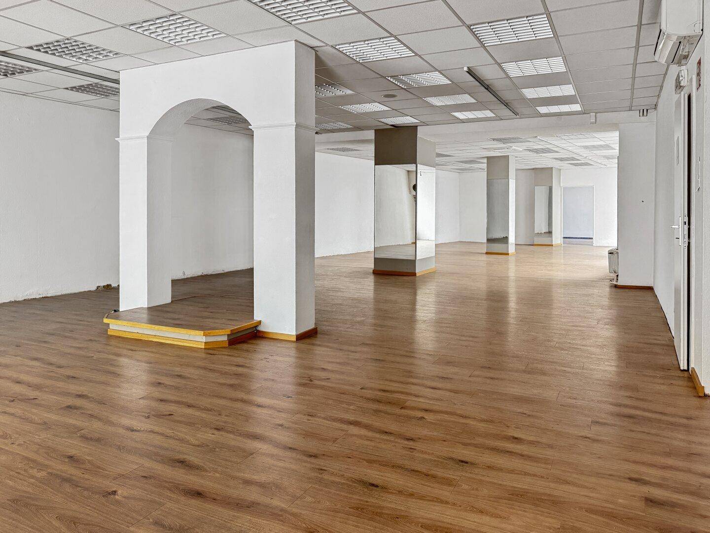 Geräumiger Gewerberaum mit Laminatboden, Rasterdecke, verspiegelten Säulen und einem Bogen.