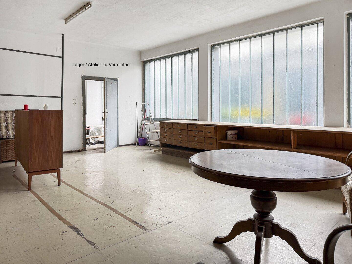 Raum mit Holzregalen, einem runden Tisch und großen Industriefenstern.