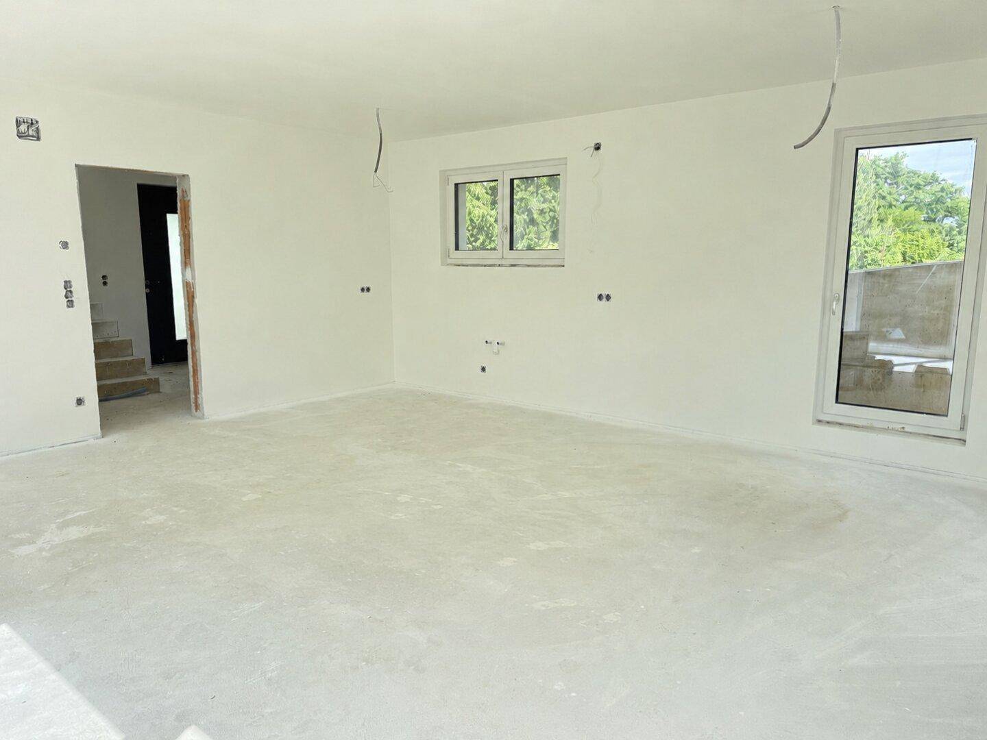 Heller, unfertiger Raum mit Betonboden, weißen Wänden und großer Glastür zum Außenbereich.