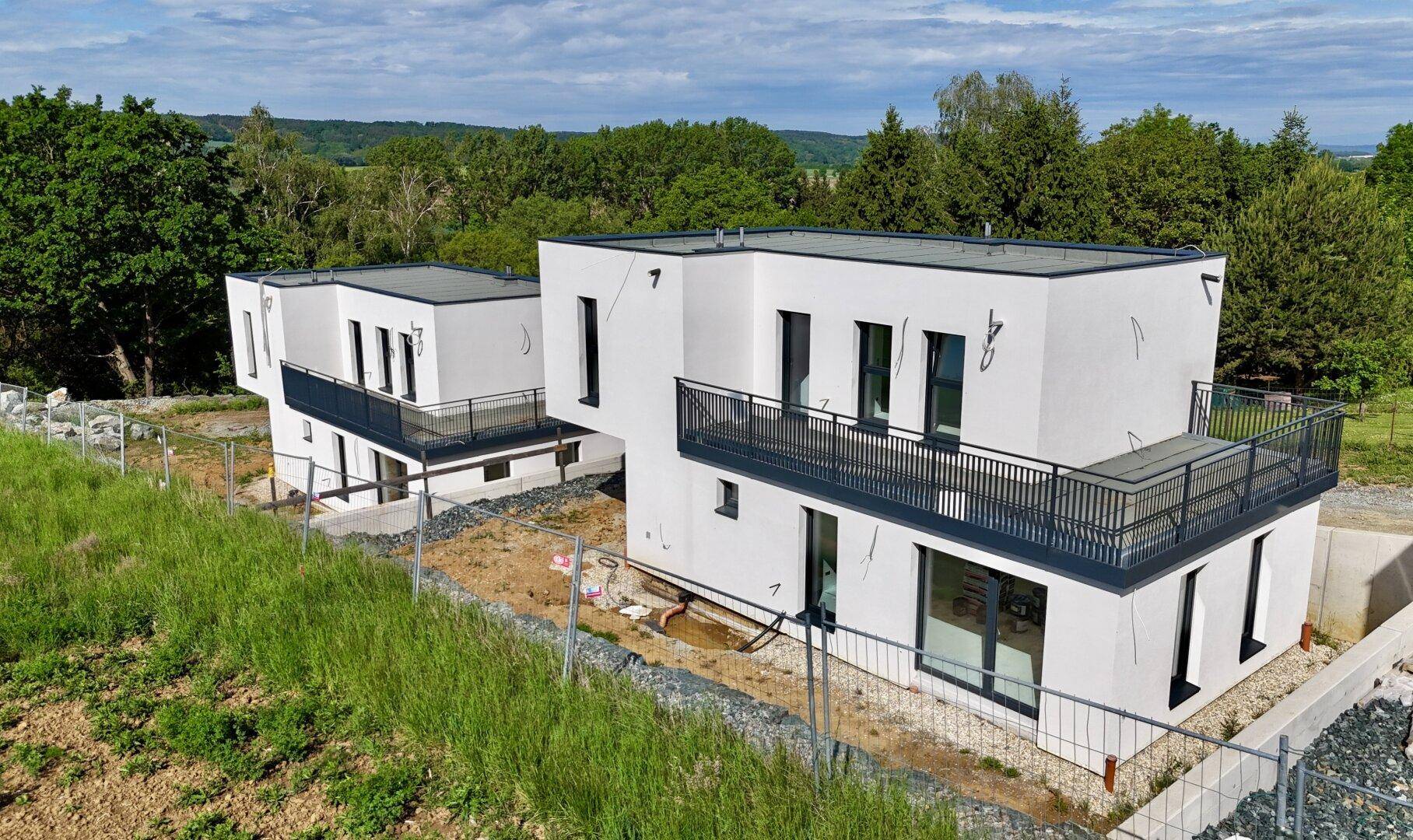 Zwei moderne weiße Häuser im Bau mit Flachdächern und Balkonen, umgeben von grüner Landschaft.