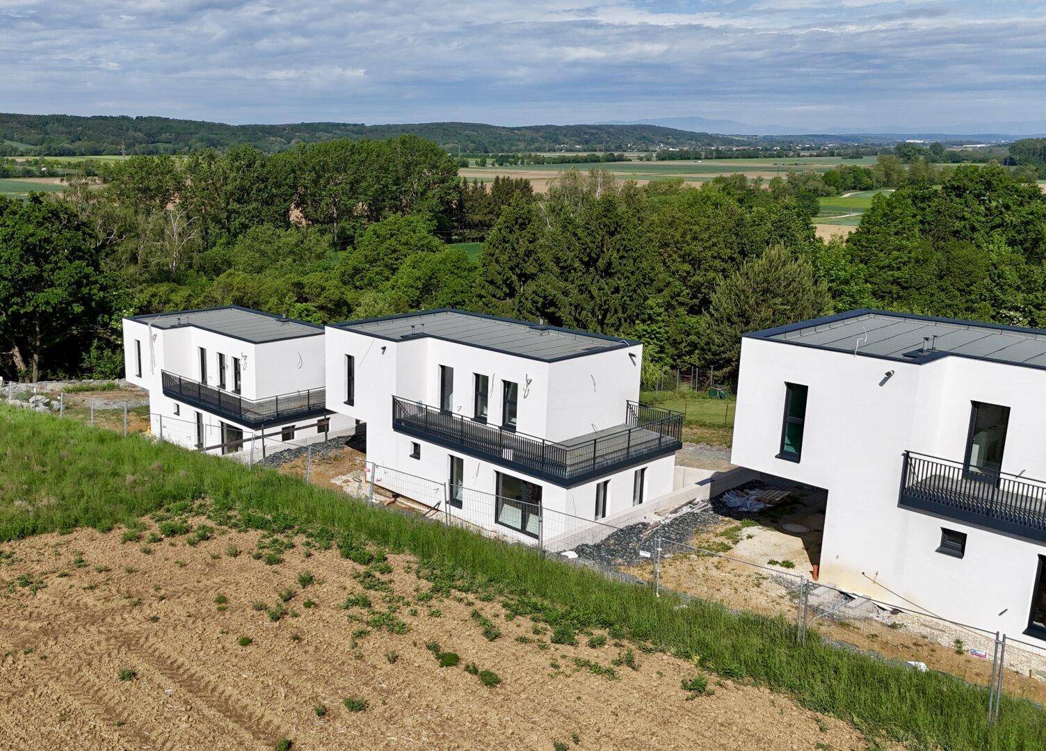 Drei moderne weiße Häuser im Bau mit Flachdächern und Balkonen, umgeben von Feldern und Wäldern.