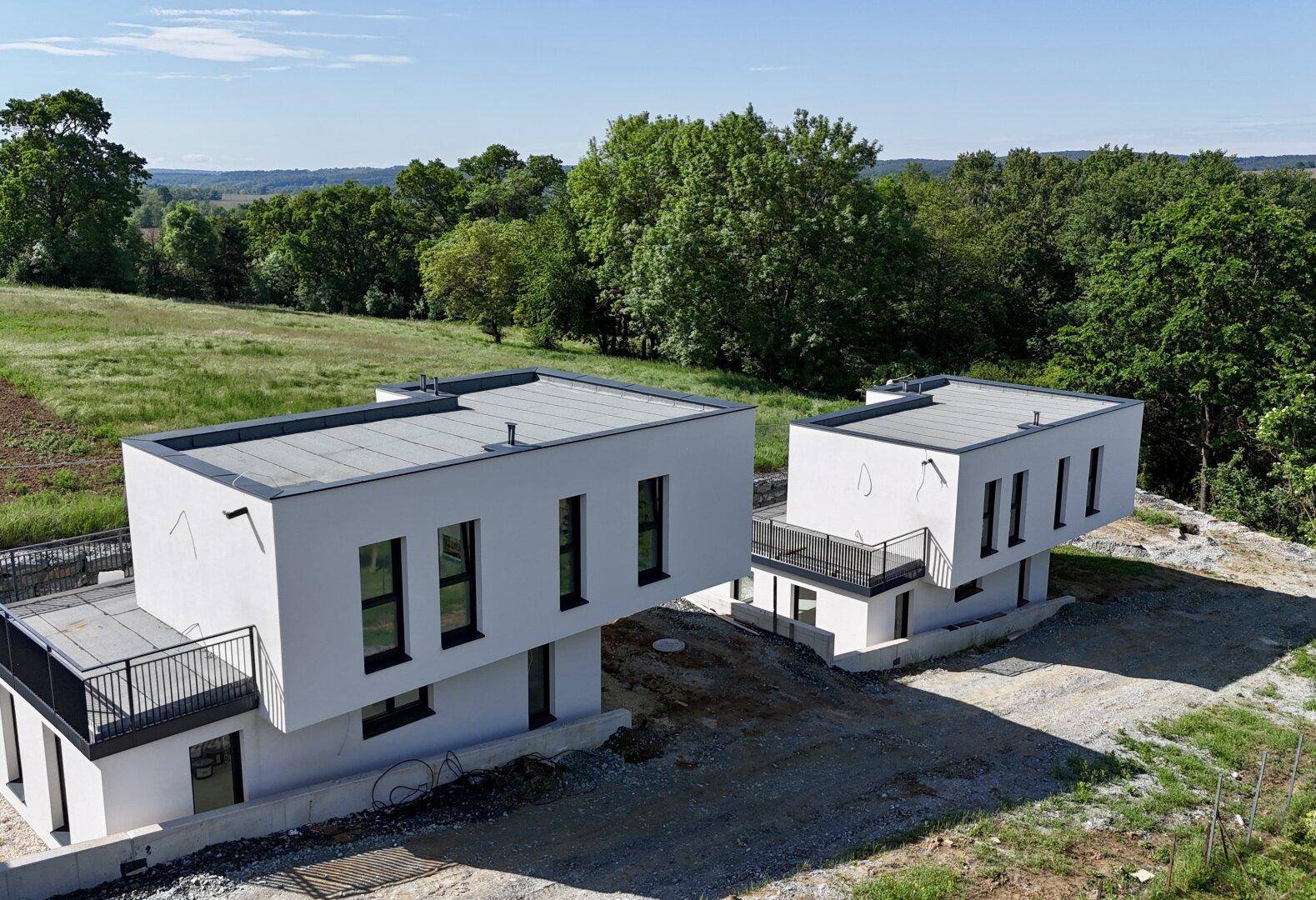 Zwei moderne Einfamilienhäuser mit Flachdach in ländlicher Umgebung unter blauem Himmel.