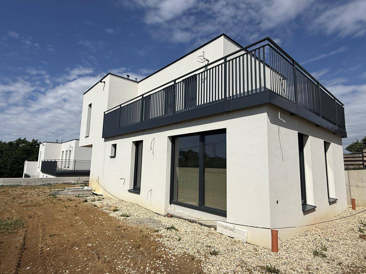 Seitenansicht eines modernen weißen Hauses mit Balkon und unentwickeltem Gelände unter blauem Himmel.
