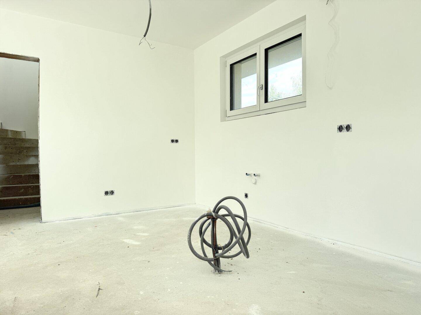 Unfertiger Raum mit Betonboden, weißen Wänden und Fenster, mit sichtbaren Elektroinstallationen.