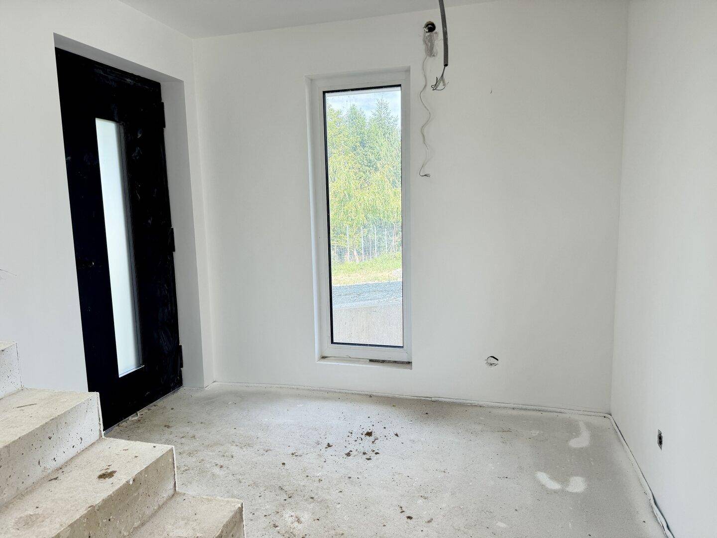 Unfertiger Eingangsbereich mit Betonboden, weißen Wänden und hohem Fenster mit Blick ins Grüne.