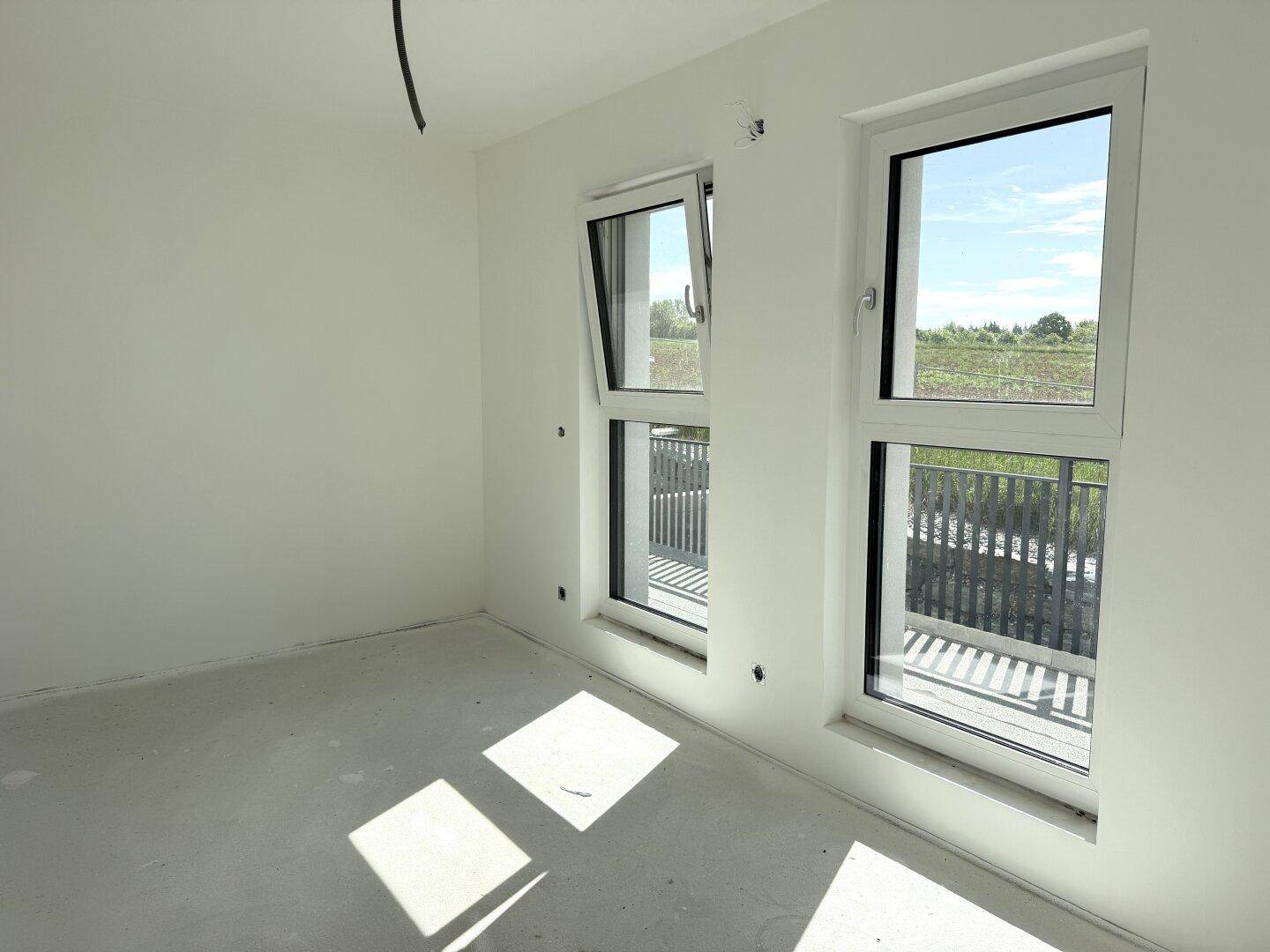 Helles, unfertiges Schlafzimmer mit Betonboden und zwei großen Fenstern, eines davon gekippt.