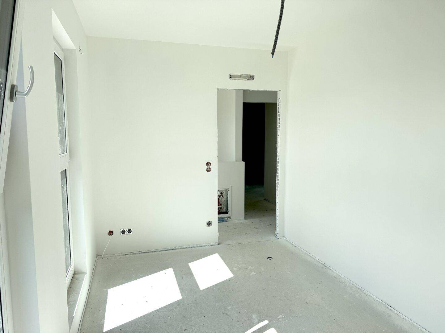 Helles, unfertiges Schlafzimmer mit Betonboden, weißen Wänden und großem Fenster.
