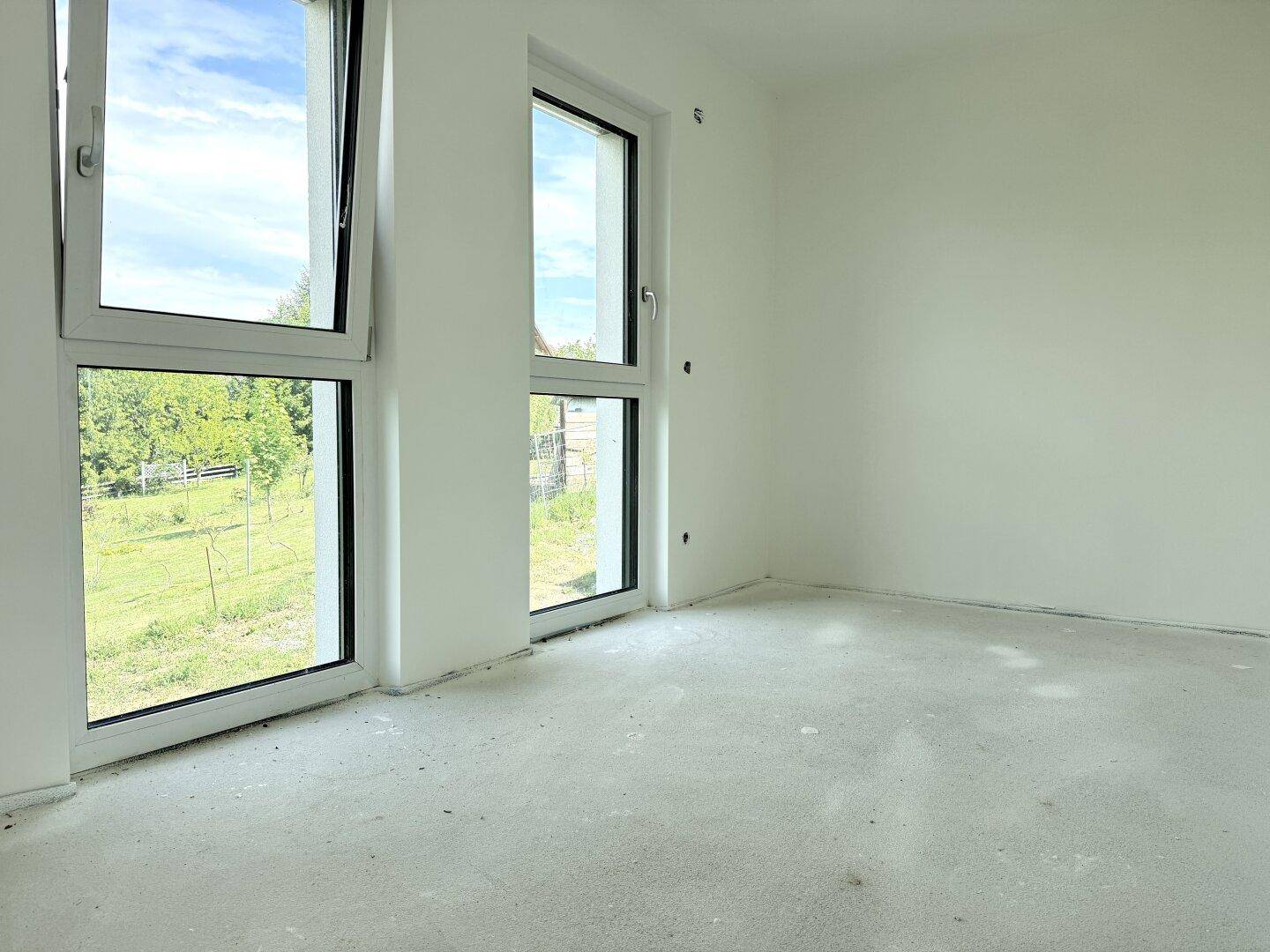 Unfertiges Schlafzimmer mit Betonboden, weißen Wänden und zwei großen Fenstern mit grüner Aussicht.
