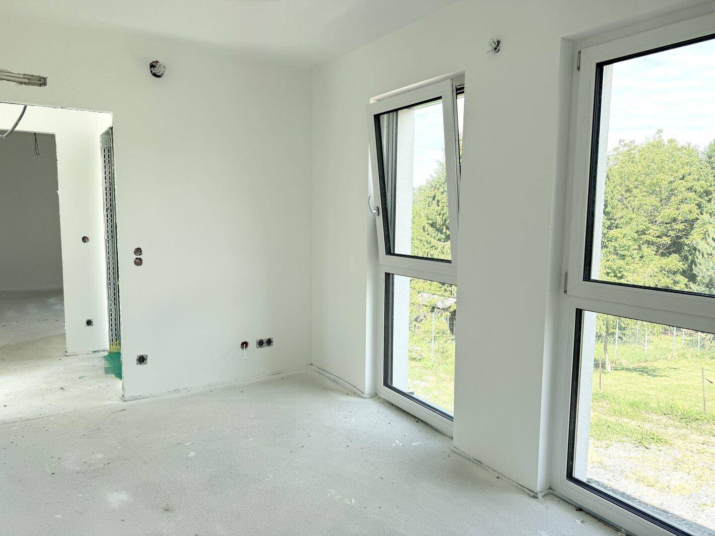 Unfertiges Schlafzimmer mit Betonboden, weißen Wänden und Fenstern mit Blick ins Grüne.
