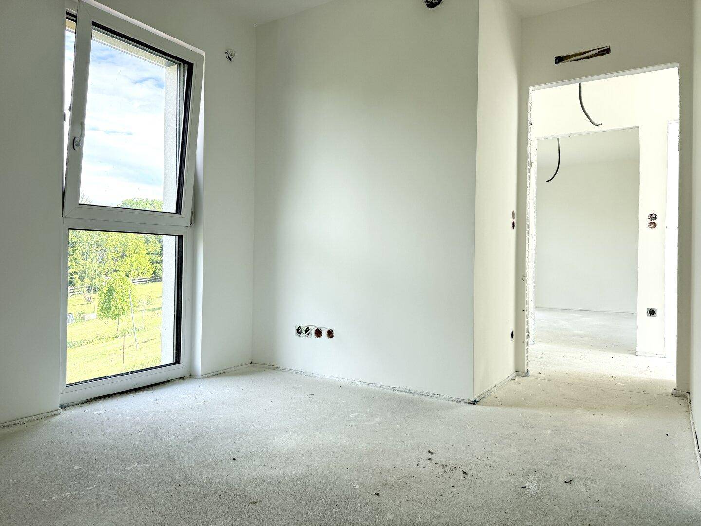 Unfertiges Schlafzimmer mit Betonboden, weißen Wänden und großem Fenster, das viel Licht hereinlässt.