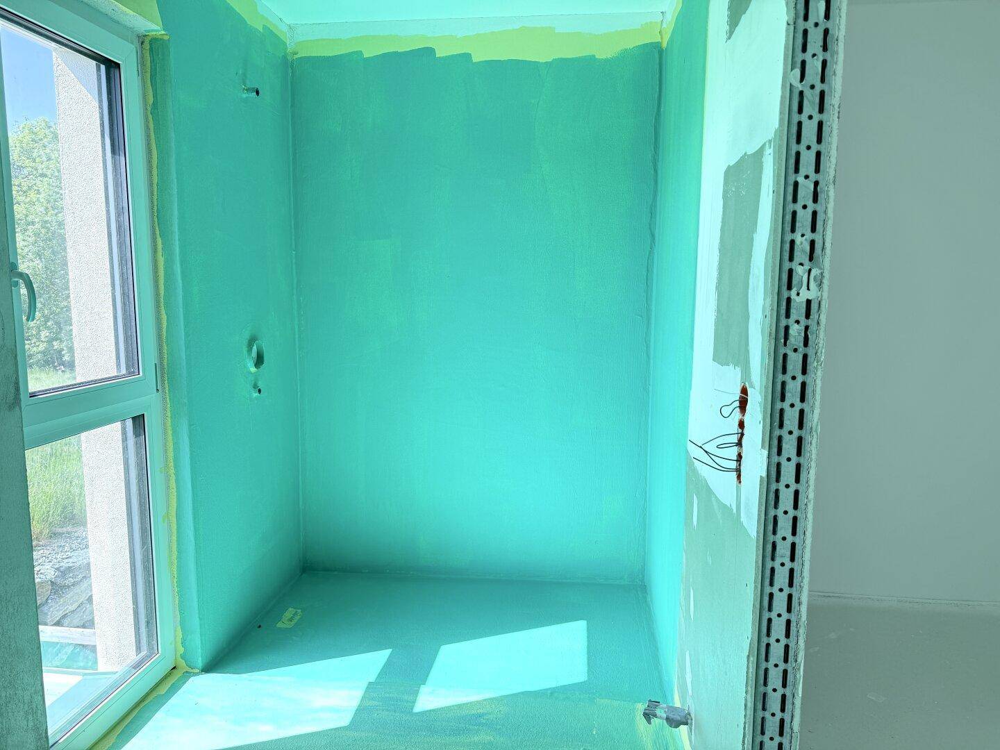 Unfertiges Badezimmer mit türkisfarbener Abdichtung an Wänden und Boden sowie einem Fenster.