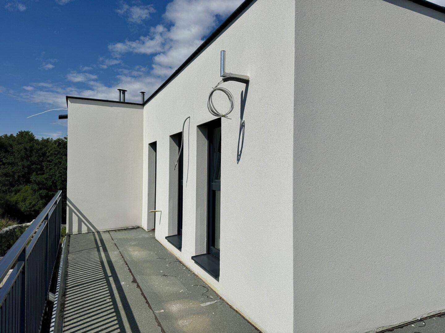Langer Balkon mit dunklem Geländer an der Seite eines modernen weißen Hauses unter blauem Himmel.